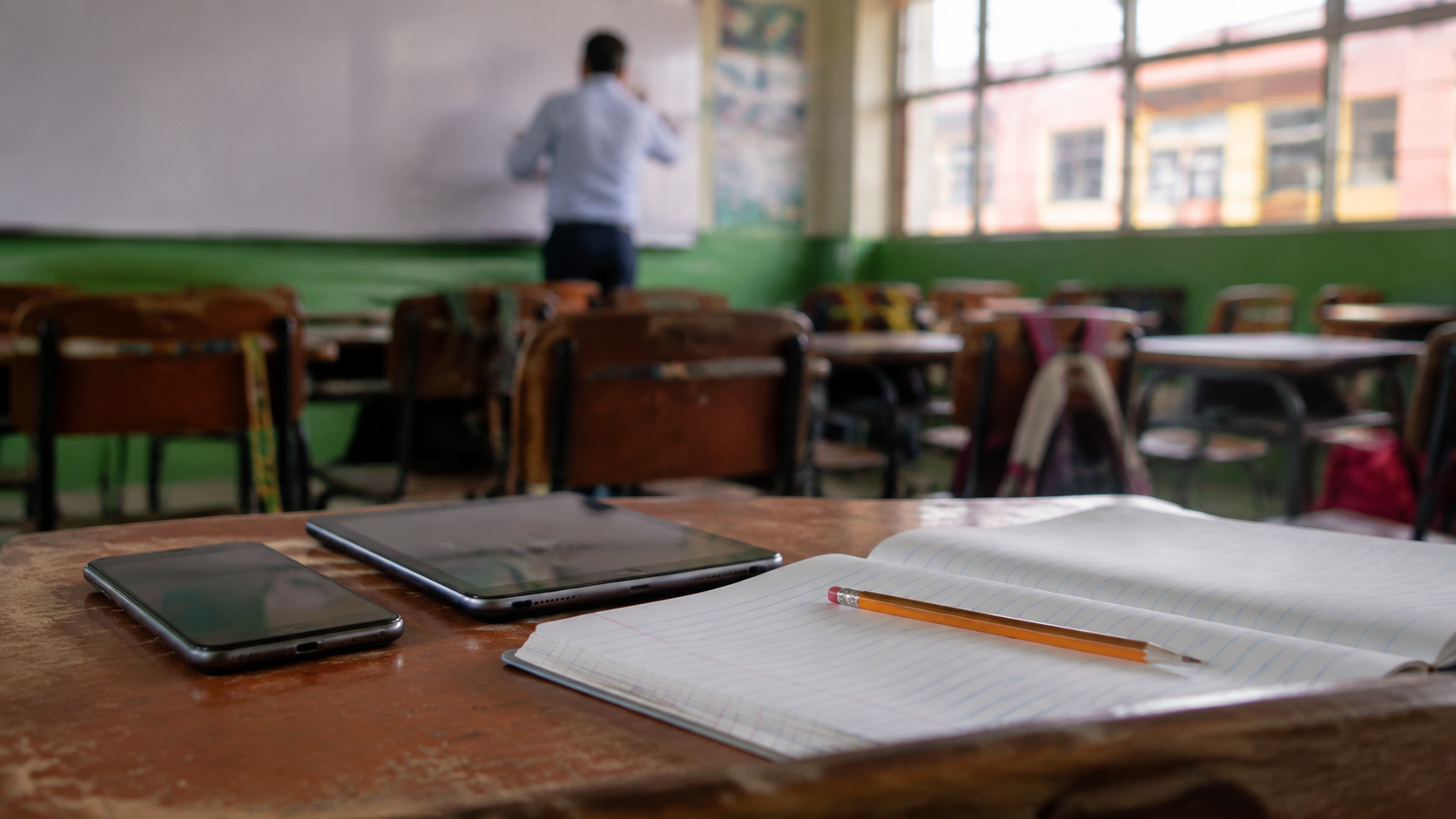 Teléfono y tableta sobre un pupitre junto a un cuaderno y lápiz dentro de un salón de clases, imagen editorial sobre dispositivos digitales en escuelas.